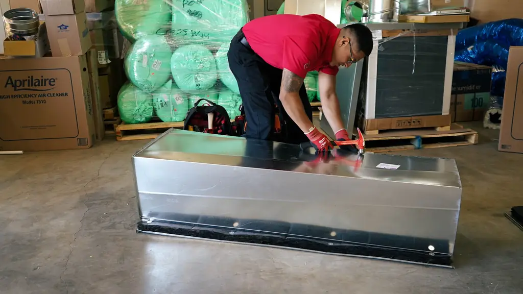 Crossway technician hammering a fabricated sheet-metal plenum on the warehouse floor, an Aprilaire Model 1510 carton behind him